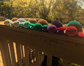 Könnte beinhalten: Eine Sammlung farbenfroher, handgefertigter Häkeltiere, darunter Schildkröten, Pilze und ein Oktopus, angeordnet auf einem Holzgeländer. Die Spielzeuge sind in verschiedenen Grüntönen, Braun, Lila und Rot gehalten und weisen sichtbare Nähte und weiche Texturen auf.