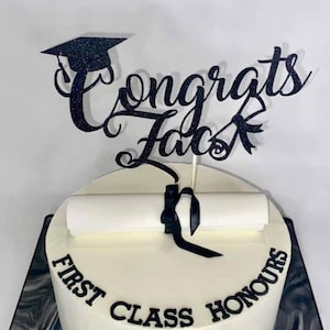 Op de afbeelding: Een ronde, wit geglazuurde taart met de woorden "FIRST CLASS HONOURS" in zwarte letters. Een zwarte afstudeerhoed en de woorden "Congrats Jack" staan erop. De taart staat op een zwart-grijze marmeren basis.