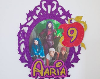 May include: Purple glitter frame with a picture of the characters from the movie Descendants. The frame has a red apple with a gold 9 inside. The name "Aria" is written in gold and pink letters below the frame.