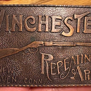 以下が含まれることがあります： 「Winchester Repeating Arms」の文字とライフルデザインが施された、長方形のブロンズ色の金属製ベルトバックル。「New Haven Conn」の文字も確認できます。