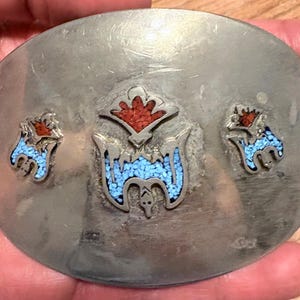 May include: Oval silver-toned belt buckle with inlaid turquoise and red accents. The buckle features three decorative elements: a central design and two smaller side designs. The central design has a bat-like shape with turquoise inlay.