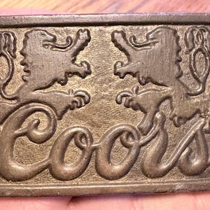 Puede incluir: Hebilla de cinturón rectangular de color bronce con dos leones heráldicos que flanquean la palabra "Coors" en una escritura estilizada. La hebilla tiene un aspecto vintage con detalles en relieve.
