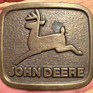 Könnte beinhalten: Rechteckige Gürtelschnalle aus Messing mit einer springenden Hirschsilhouette. Die Worte "JOHN DEERE" sind unter dem Hirsch geprägt. Die Schnalle hat eine strukturierte Oberfläche und ein Vintage-Aussehen.