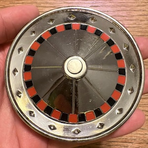 May include: A circular silver-toned metal belt buckle featuring a central raised disc surrounded by a ring of alternating red and black enamel squares. The outer rim is decorated with small, evenly spaced pyramid-shaped studs in a silver finish.