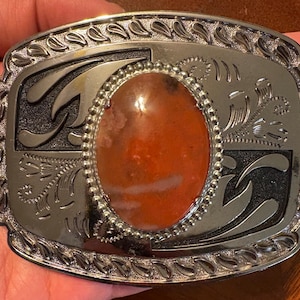 Puede incluir: Hebilla de cinturón de metal plateado con una piedra ovalada de color rojo anaranjado en el centro. La hebilla tiene un borde decorativo y diseños florales y de volutas grabados. La piedra está rodeada por un marco plateado con cuentas.