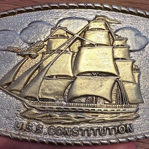 May include: A rectangular gold-tone metal belt buckle with a rope-style border. The center features a raised relief design of the U.S.S. Constitution sailing ship with an eagle flying above it. The text U.S.S. CONSTITUTION is embossed at the bottom.