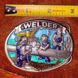 May include: Oval metal belt buckle with the word "WELDER" at the top. The buckle features a colorful scene of welders working on a pipe, with a crane in the background. The buckle is approximately 3 inches long.