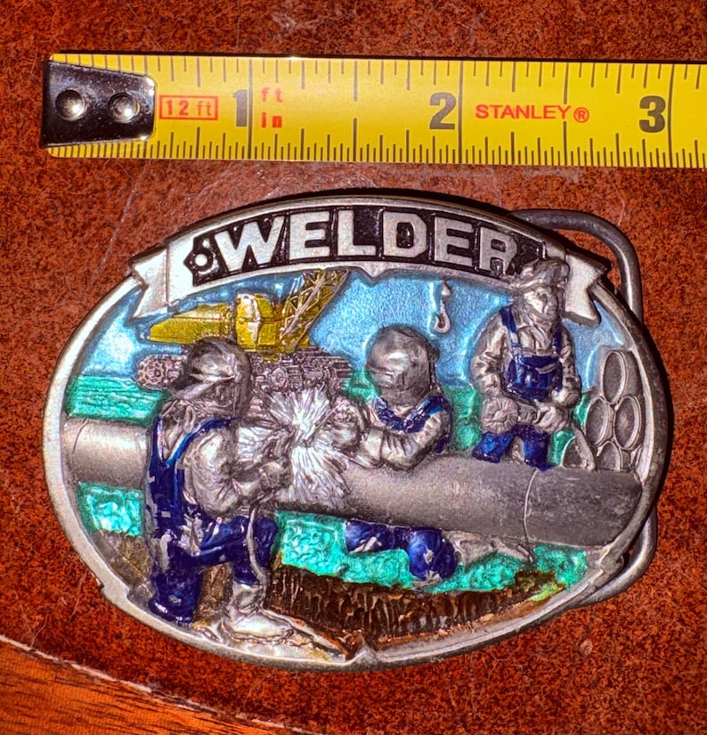 May include: Oval metal belt buckle with the word "WELDER" at the top. The buckle features a colorful scene of welders working on a pipe, with a crane in the background. The buckle is approximately 3 inches long.