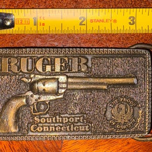 以下が含まれることがあります： ヴィンテージ真鍮ベルトバックル。リボルバーのレリーフと「RUGER」の文字が特徴です。「Southport, Connecticut」と「THE PROOF OF QUALITY」の文字も含まれています。バックルの長さは約7.6cmです。