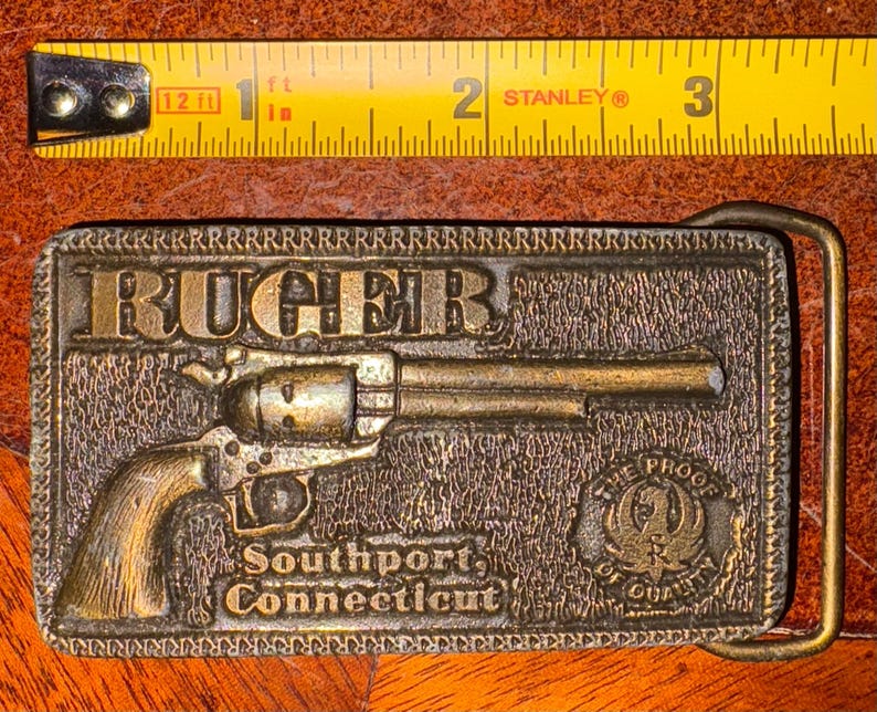 以下が含まれることがあります： ヴィンテージ真鍮ベルトバックル。リボルバーのレリーフと「RUGER」の文字が特徴です。「Southport, Connecticut」と「THE PROOF OF QUALITY」の文字も含まれています。バックルの長さは約7.6cmです。