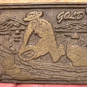 Peut inclure: Boucle de ceinture rectangulaire en métal sur le thème de l'exploitation minière de l'or. La boucle présente un relief d'un chercheur d'or, avec le mot "GOLD" au-dessus. Le design comprend des montagnes et de l'eau.