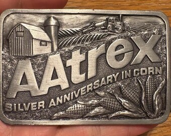 May include: Silver-colored belt buckle with the word "AAtrex" in large letters, with the text "Silver Anniversary in Corn". The buckle features a barn, silo, tractor, and corn stalks.