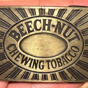 Può includere: Fibbia per cintura rettangolare in ottone con la scritta in rilievo "BEECH-NUT CHEWING TOBACCO". Il testo è all'interno di una forma ovale, circondata da un disegno a raggi solari. La fibbia ha un aspetto vintage.