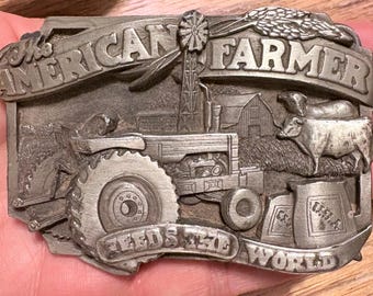 Op de afbeelding: Zilverkleurige metalen riemgesp met de woorden "The American Farmer" en "Feeds the World". De gesp toont een tractor, schuur, windmolen en vee.