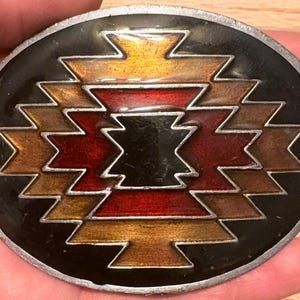 May include: Oval belt buckle with a black background and a geometric design in shades of gold, brown, and red. The buckle has a silver-coloured metal border. The design is a zig-zag pattern.