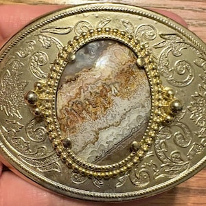 May include: An oval-shaped gold-tone belt buckle featuring an intricate embossed floral scrollwork pattern across the surface. The center contains an oval inlay of natural stone with swirling brown, white, and gray patterns, framed by a beaded border.