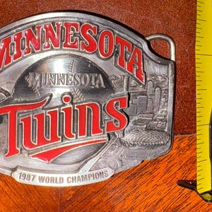 May include: A silver-colored belt buckle with the words "MINNESOTA" and "Twins" in red lettering. The buckle features a baseball and a cityscape design, with the text "1987 WORLD CHAMPIONS".