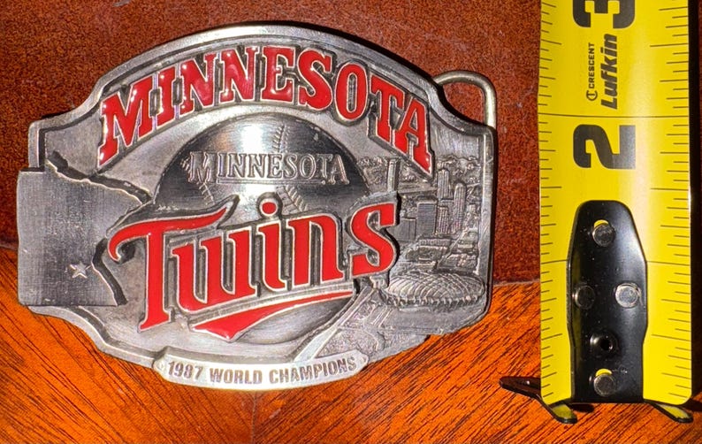 May include: A silver-colored belt buckle with the words "MINNESOTA" and "Twins" in red lettering. The buckle features a baseball and a cityscape design, with the text "1987 WORLD CHAMPIONS".