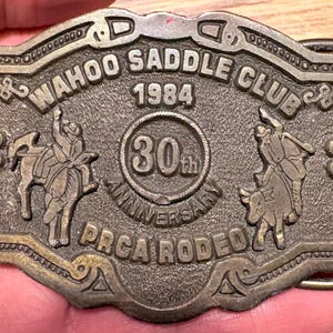 Op de afbeelding: Een vintage bronzen gesp met de tekst "WAHOO SADDLE CLUB 1984" en "30th ANNIVERSARY PRCA RODEO". De gesp heeft afbeeldingen van een ruiter op een paard en een ruiter op een stier.