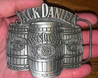 May include: A silver-colored metal belt buckle featuring three barrels with the text "JACK DANIELS" and "Old No. 7 BRAND". The buckle has a rectangular shape with a curved bar for the belt.