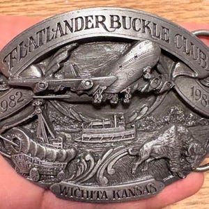 May include: An oval-shaped pewter belt buckle featuring the text FLATLANDER BUCKLE CLUB, 1982, 1989, and WICHITA KANSAS. The relief design depicts an airplane in flight above a riverboat, a covered wagon, and a buffalo, all rendered in metallic gray.