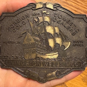 May include: An antique brass belt buckle with a detailed relief of a sailing ship. The buckle is rectangular with curved edges and features the text "FOREIGN AND GOLD DOMESTIC DEALERS" and "LIVINGSTON WELLS & CO". The buckle also has the text "AUSTRALIA" and "SOUTH AFRICA".