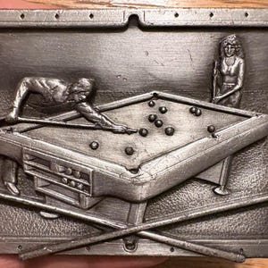May include: A rectangular silver-toned metal belt buckle featuring a raised relief design of a pool table with a person leaning over to take a shot. A second person stands nearby holding a cue, and two crossed pool cues are depicted at the bottom.