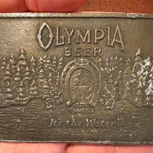 Puede incluir: Hebilla de cinturón rectangular de color bronce con el logotipo de Olympia Beer. El diseño incluye el nombre de la marca, una herradura, una escena de bosque y la frase "It's the Water".