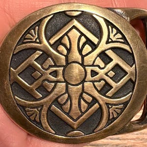 May include: A round, antique brass belt buckle with an intricate, symmetrical design. The central motif is a cross-like shape with ornate detailing, surrounded by a circular border. The buckle has a rectangular bar for the belt.