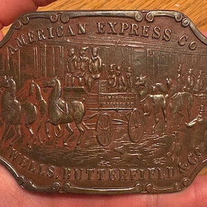 Puede incluir: Una hebilla de cinturón metálica rectangular vintage con un diseño en relieve. La hebilla presenta el texto "AMERICAN EXPRESS CO" y "WELLS, BUTTERFIELD & CO" alrededor de una escena detallada de un carruaje tirado por caballos y figuras. La hebilla es de color bronce.