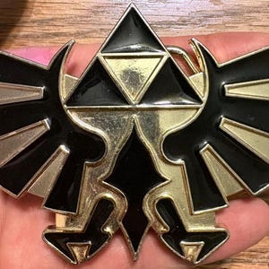 Könnte beinhalten: Eine Gürtelschnalle aus goldfarbenem und schwarzem Metall mit einem stilisierten Vogel-Emblem und einem zentralen dreieckigen Design. Das Stück hat ein poliertes Gold-Finish mit schwarzer Emaille-Einlage an den Flügeln und der Mitte.