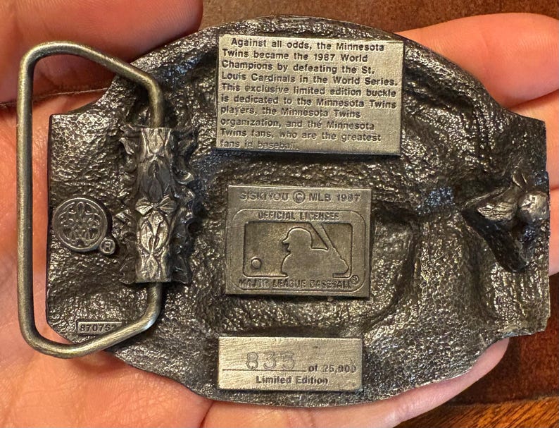 May include: A detailed, silver-toned metal belt buckle commemorating the Minnesota Twins' 1987 World Series victory. The buckle features text detailing the team's achievement, the MLB logo, and the limited edition number 833 of 25,000.