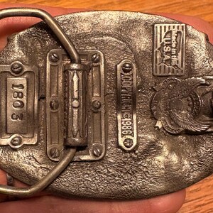 May include: Vintage metal belt buckle with an oval shape. The buckle features the text "1803" and "COPYRIGHT 1986". It also has an emblem with an eagle and the text "Made in the U.S.A."