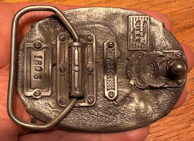 May include: Vintage metal belt buckle with an oval shape. The buckle features the text "1803" and "COPYRIGHT 1986". It also has an emblem with an eagle and the text "Made in the U.S.A."