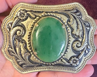 May include: Rectangular belt buckle with a large oval green stone center. The buckle has a gold-tone frame with ornate silver-tone floral and scroll designs. The edges are detailed with small, raised beads.