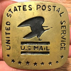May include: A square brass belt buckle with rounded corners featuring the United States Postal Service logo. The design includes an embossed eagle silhouette, the text U.S. MAIL, and a border of stars along the bottom edge of the metal surface.