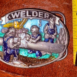 May include: Oval metal belt buckle with the word WELDER at the top. The buckle features a colorful scene of three welders working on a pipe, with a crane in the background. The buckle is silver with blue, green, and yellow accents.
