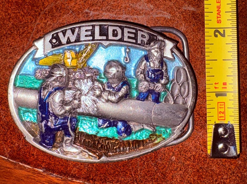 May include: Oval metal belt buckle with the word WELDER at the top. The buckle features a colorful scene of three welders working on a pipe, with a crane in the background. The buckle is silver with blue, green, and yellow accents.