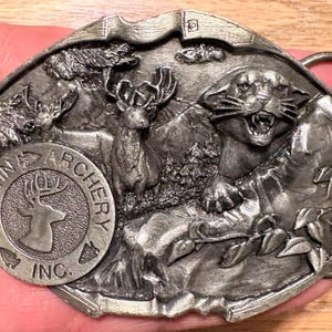 Puede incluir: Hebilla de cinturón de metal plateado con un diseño en relieve detallado. La hebilla muestra una escena de montaña con un ciervo, un puma gruñendo y el texto "MARTIN ARCHERY INC." en un emblema circular. La hebilla tiene forma rectangular con bordes redondeados.