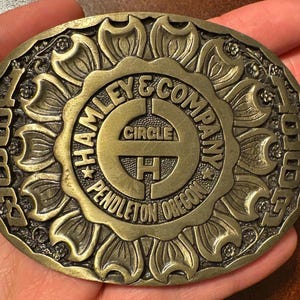May include: A round, antique brass belt buckle with a floral design around the edge. The center features the text "HAMLEY & COMPANY CIRCLE H PENDLETON OREGON". The buckle is held in a hand.