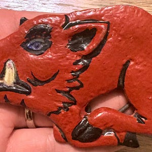 May include: A red, flat, shaped object depicting a wild boar in profile. The boar has black painted outlines, a single visible white tusk, and a small purple eye. The surface has a textured, glossy finish with a cutout section near the legs.