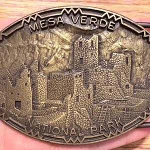 May include: An oval-shaped, bronze-colored belt buckle featuring a detailed relief of Mesa Verde National Park. The buckle has the words "MESA VERDE" and "NATIONAL PARK" engraved on it. The design includes depictions of ancient cliff dwellings and mountainous terrain.