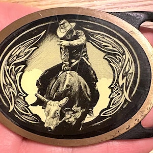 May include: Oval-shaped belt buckle with a cowboy riding a bull, set against a yellow background. The buckle has a black frame with a gold-colored border and a black loop for the belt.