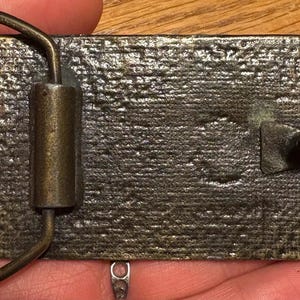 以下が含まれることがあります： 長方形のアンティークスタイルのベルトバックル。表面はテクスチャ加工されたブロンズ色です。バックルには、ベルトを固定するためのループとピン機構があります。金属は使い古された外観で、古さやヴィンテージスタイルを示唆しています。