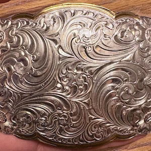 Puede incluir: Hebilla de cinturón plateada con un diseño floral y de volutas ornamentado. La hebilla rectangular tiene un borde dorado y presenta patrones detallados en relieve. El diseño incluye flores y hojas arremolinadas, creando una estética vintage clásica.