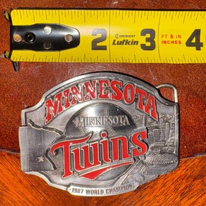 May include: A silver-colored metal belt buckle featuring the words "MINNESOTA" and "Twins" in red lettering. The buckle also includes the text "1987 WORLD CHAMPIONS" and a baseball design. A yellow tape measure is in the background.