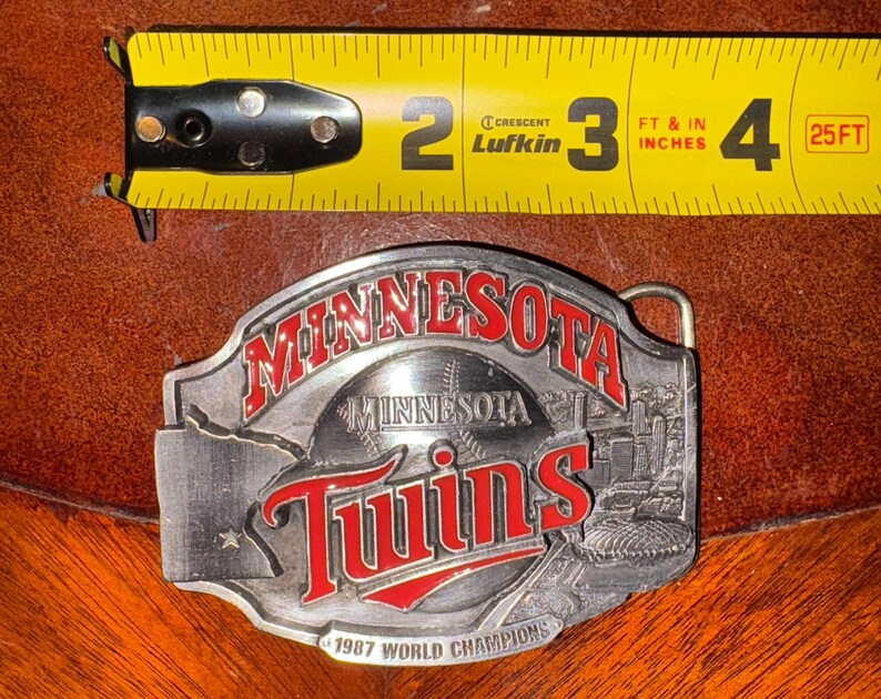 May include: A silver-colored metal belt buckle featuring the words "MINNESOTA" and "Twins" in red lettering. The buckle also includes the text "1987 WORLD CHAMPIONS" and a baseball design. A yellow tape measure is in the background.