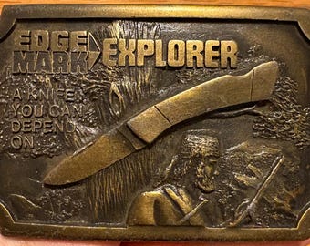 May include: A rectangular, bronze-coloured belt buckle with the words "EDGE MARK EXPLORER" and "A KNIFE YOU CAN DEPEND ON" embossed on it. The buckle features a knife and a detailed relief of a person in a wilderness setting.