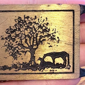 May include: Rectangular brass belt buckle with a black tree and animal silhouette design. The buckle has a black frame and a single bar for the belt. The tree is detailed with many branches and leaves, and the animals are grazing under the tree.
