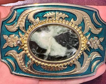 May include: An ornate belt buckle with a teal enamel background and gold-colored filigree. The center features an oval stone with green and white marbling. Floral and leaf designs adorn the edges, creating a vintage aesthetic.